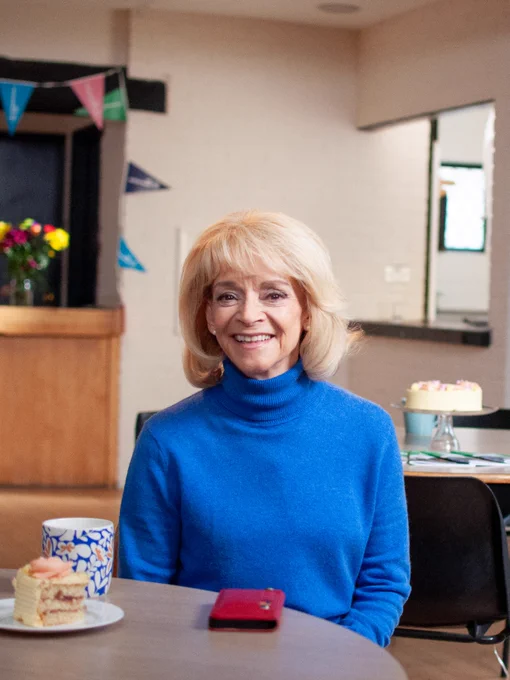 Actor Sue Holderness is sat in a cafe wearing a bright blue roll-neck jumper. On the table in front of her is a mobile, a mug of tea and a slice of cake. In the background the rest of the cake can be seen. She smiles warmly at the camera.