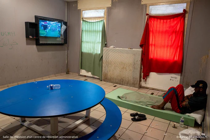 Dans la salle de télévision d'un centre de rétention administrative, un homme assis sur un matelas posé au sol regarde la TV fixée au mur. Une table ronde se trouve au centre de la pièce qui est sale et dégradée (traces de salissures et graffitis sur les murs, papiers au sol). Des couvertures sont accrochées aux fenêtres comme des rideaux de fortune -  crédit photo : Karine Bizard / CGLPL
