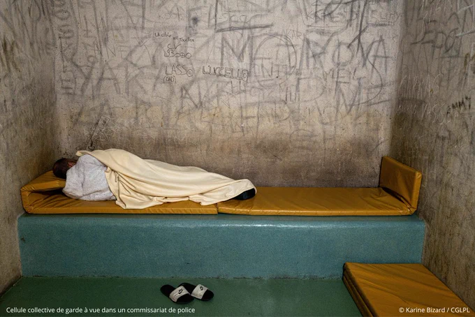 Cellule collective de garde à vue dans un commissariat de police. Un homme est allongé sur le bat-flanc de la cellule, ses claquettes sont posées par terre. Il est de dos et recouvert d'une couverture. Des matelas sont disposés sur le bat-flanc et sur le sol de la cellule. Les murs sont recouverts d'inscriptions et de graffitis - crédit photo : Karine Bizard / CGLPL