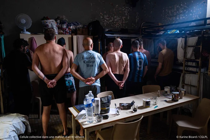 Cellule collective dans une maison d'arrêt vétuste. Au sein d’une cellule sombre, 9 hommes se tiennent debout, de dos. Des armoires et deux lits superposés à 3 étages se trouvent au fond de la pièce contre le mur. Au centre de la pièce il y a une grande table entourée de 4 chaises - crédit photo : Karine Bizard / CGLPL
