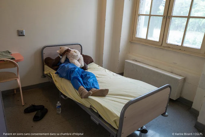 Patient dans sa chambre d'hôpital psychiatrique. Un patient en pyjama bleu d'hôpital est allongé sur le lit de sa chambre, il tient dans des bras un gros ours en peluche - crédit photo : Karine Bizard / CGLPL