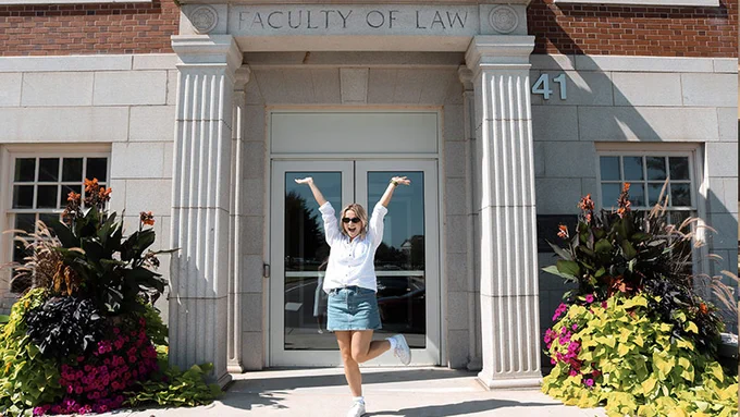 UNB graduate Hayley Bone stands joyfully with arms raised in front of a building entrance labeled "Faculty of Law." She is surrounded by vibrant flower arrangements. 