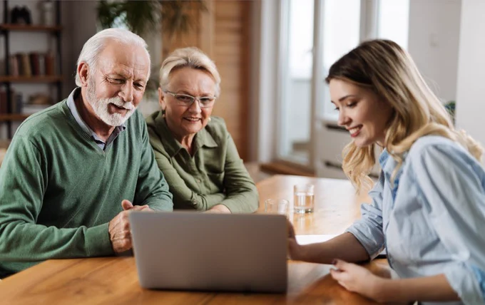 Senior couple attentively listening to a woman explaining something on a laptop at a wooden table in a bright room.