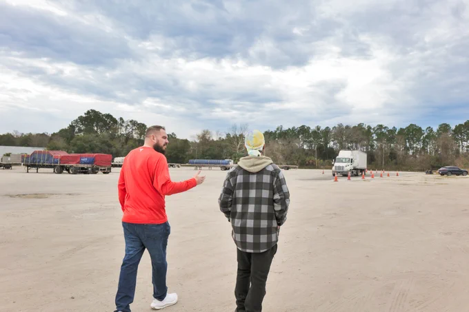 Two men walking across a large CDL training yard with trucks and trailers in the background under a cloudy sky.