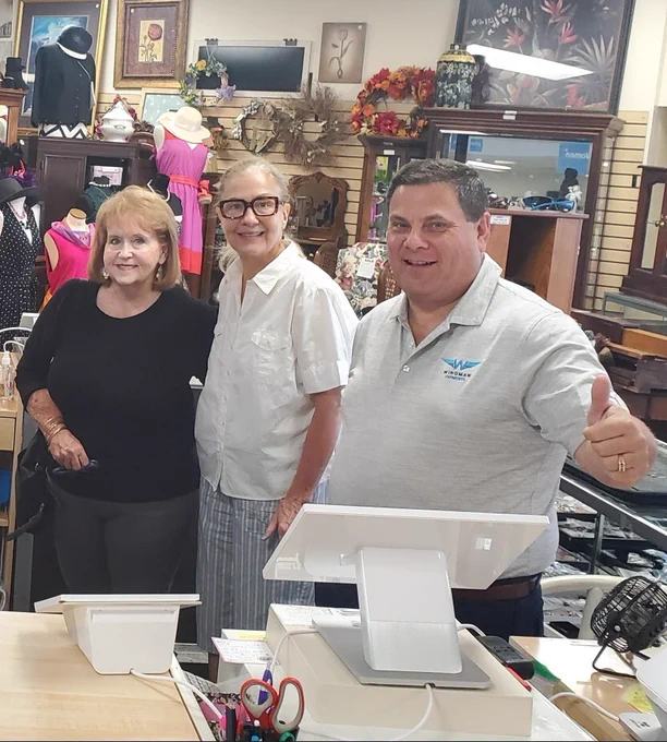 Group photo at a retail checkout counter. Seven staff or volunteers smile behind the register area while a Wingman Payments representative gives a thumbs up beside two white POS terminals. The shop is filled with clothing, decor, and display cases in the background.