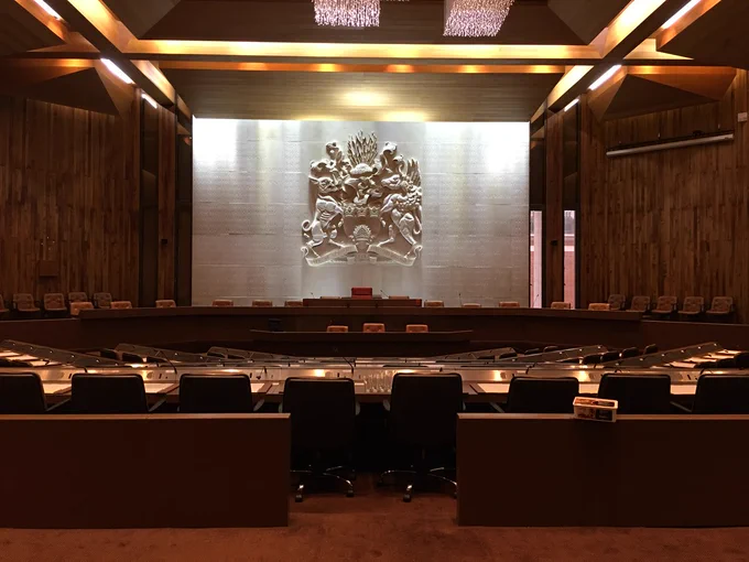 Formal chamber with wood panelling, a large crest on the back wall, and rows of desks with microphones.