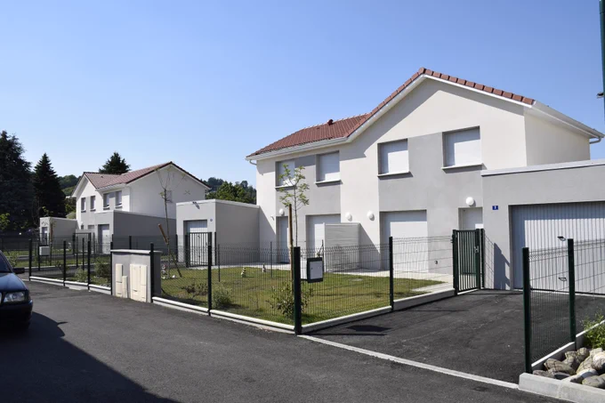 Alignement de maisons neuves à étage avec façades blanches et grises de la résidence, petits jardins privatifs clôturés et garages attenants, sous un ciel bleu.