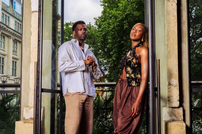 A photo showing two performers leaning against masonry looking out over a leafy quad
