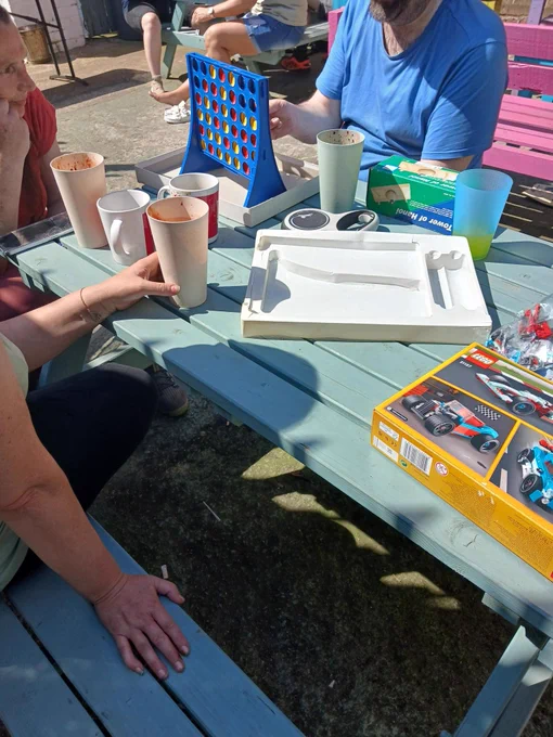 People sitting around a picnic table playing Connect Four with drinks and toy boxes scattered on the table outdoors.