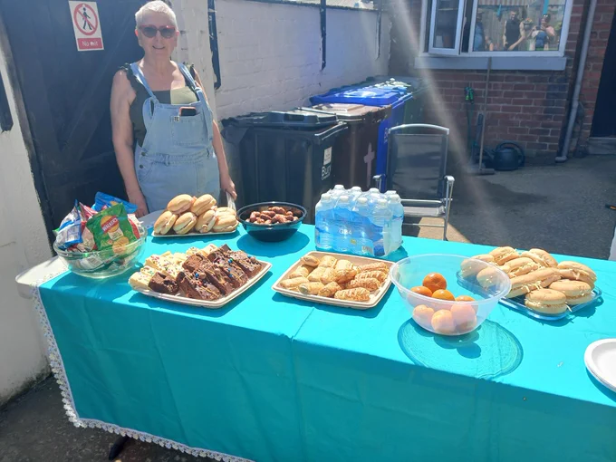 Outdoor table covered with a blue cloth displaying sandwiches, brownies, pastries, bottled water, and fruit bowls next to a person in denim overalls.