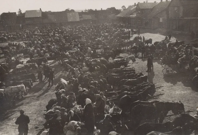 Black-and-white historical photograph taken from an elevated vantage point, showing a large town square densely packed with hundreds of people, horse-drawn carts, and bundled belongings. The crowd fills the entire square, with low-roofed buildings visible along the edges. The photograph documents the deportation operation in Uchanie on 10 June 1942, during which the Jewish population of the town was gathered in the market square before being sent to a death camp.