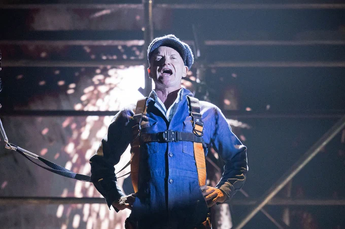 A photo showing a man in overalls shouting on a stage, next to sparks 