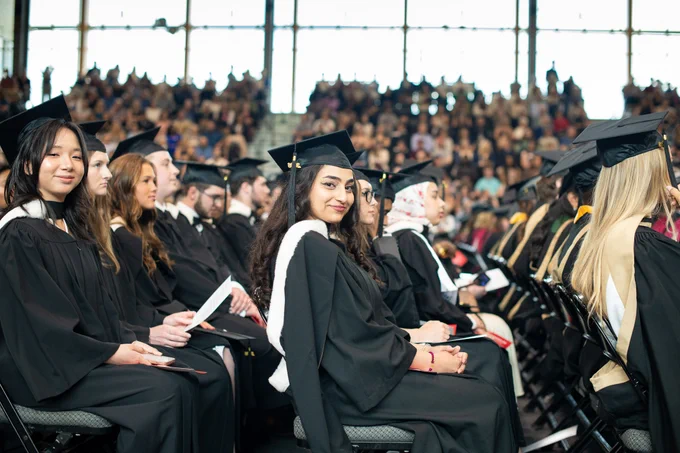A group of graduates in caps and gowns seated in rows during a commencement ceremony, with a large audience in the background.