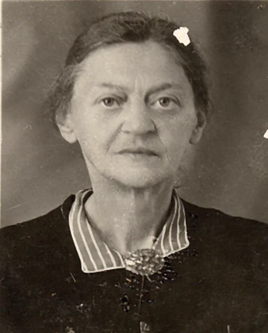 Black-and-white close-up portrait photograph of a middle-aged woman with dark hair pulled back, wearing a small hair clip. She is dressed in a dark top over a striped collared shirt, with a brooch at the collar. She looks directly at the camera with a serious expression. The photograph has the style and format of an early 20th-century identity or document photograph.