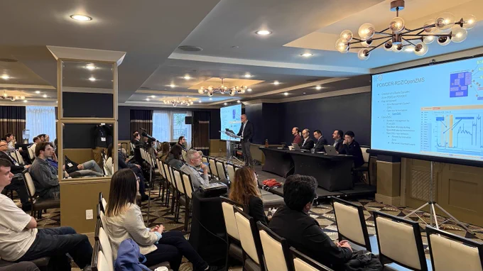 Photograph of POWDER Director Kobus Van der Merwe speaking as part of a panel at the IEEE DySPAN 2026 conference. The photograph is from the side of the presentation room. Kobus is standing at a lectern. Five other panelists are seated at a table, facing the audience.