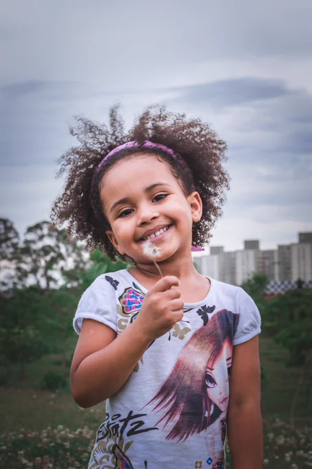 African American girl smiling at the camera with a pink headband and blowing on a dandelion.