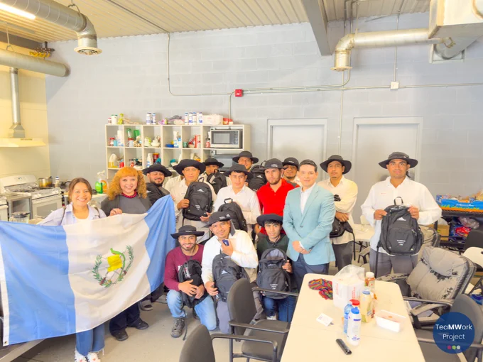 Group of workers and partners inside a facility kitchen area posing with TeaMWork backpacks, with a Guatemalan flag held at the front. Shelving with supplies, a stove, and tables are visible in the background.
