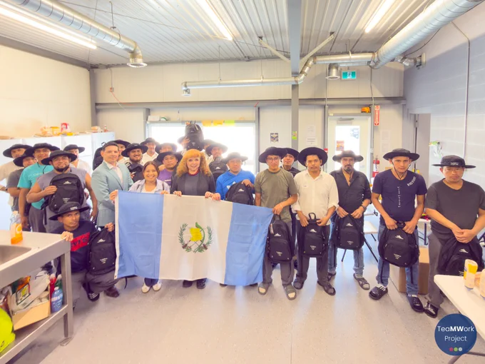 Foto de grupo tomada en el interior, en la que aparecen trabajadores extranjeros temporales y sus parejas reunidos en una sala de descanso, sosteniendo una bandera de Guatemala y mochilas de TeaMWork. En la sala se ven mesas, estanterías y alimentos mientras se reparten los kits.