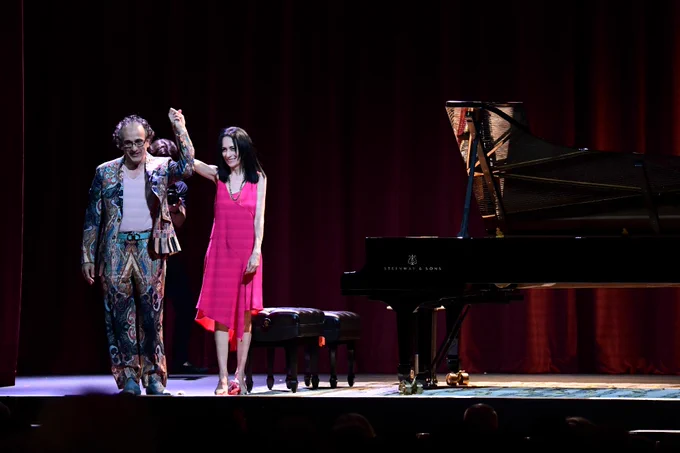 Two performers stand on stage holding hands. One is wearing a colorful patterned outfit, and the other is in a bright pink dress. A Steinway & Sons grand piano is visible nearby.