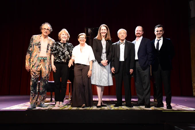 A group of seven individuals stands on a stage with a dark red curtain in the background. They are dressed in a mix of formal and colorful attire, standing side by side and smiling.