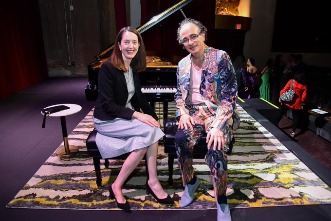 Two individuals sit on a bench in front of a grand piano. The person on the left is wearing a dark jacket and light-colored skirt, while the person on the right is dressed in a colorful, patterned suit. They are on a stage with a patterned rug underneath.