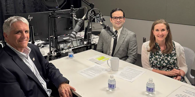 Three people sit around a table in a recording studio, with microphones, papers, and water bottles on the table.
