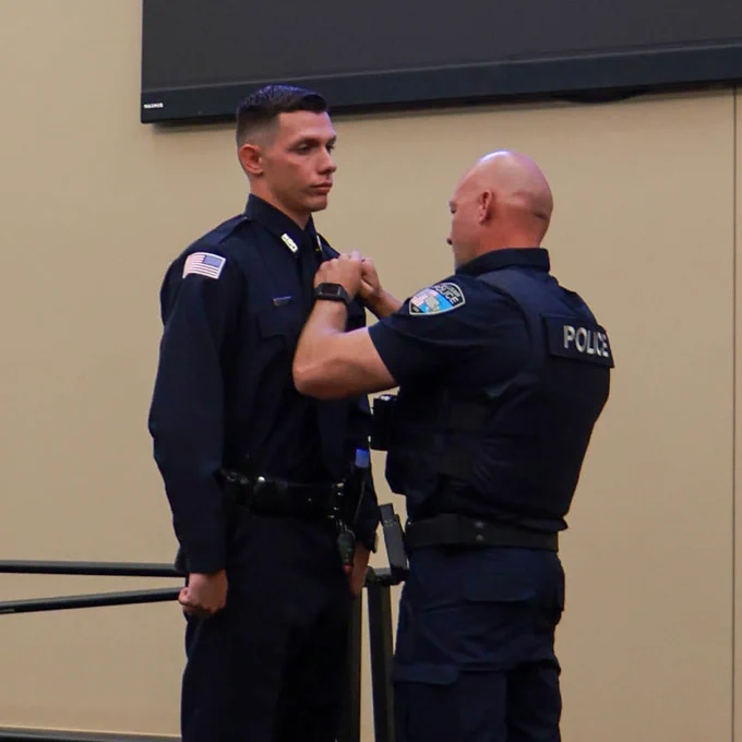 Police officer pins badge on a young officer's uniform during a formal ceremony indoors.