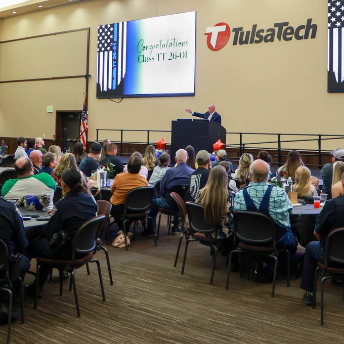 Audience seated at tables listening to a speaker congratulating Tulsa Tech Class TT 26-01 in a large event hall.