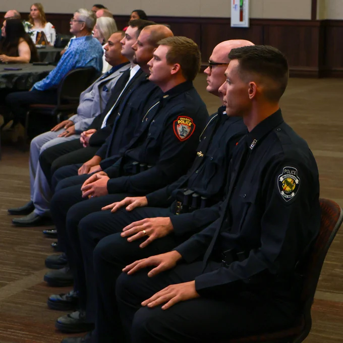 A row of police officers in uniform seated attentively during a formal indoor event with other attendees in the background.