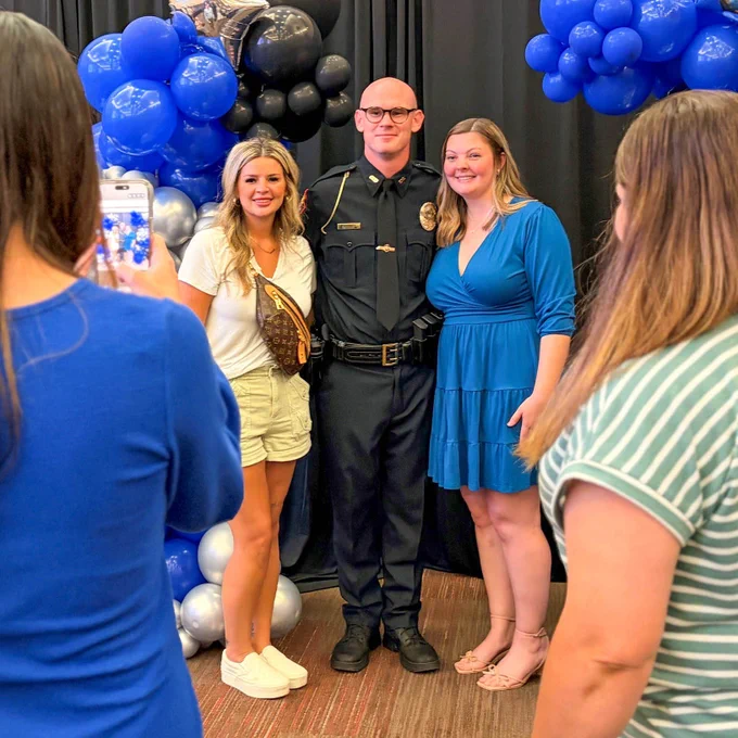 A police officer in uniform poses for a photo with two women in front of blue and black balloon decorations while others take pictures.