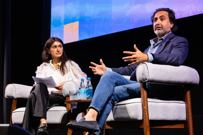 Two people on stage, moderator Ayesha Arora-Hughes (Alum) and panellist Hemant Taneja (CEO, General Catalyst) Taneja gestures with both hands while speaking. They are seated in gray armchairs, with water bottles and glasses on a small table between them. A blue and floral patterned screen is in the background.
