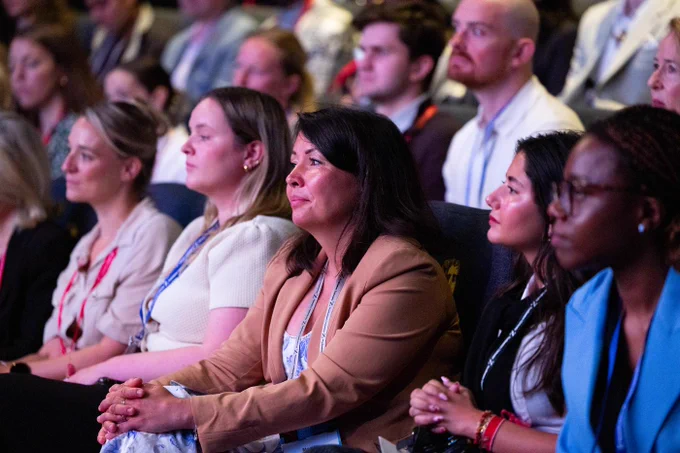 A diverse audience sits in rows at a conference, looking forward. In the foreground, a woman with dark hair, wearing a brown blazer over a white and blue floral top, clasps her hands. Other attendees, including men and women of various ethnicities, are visible, many wearing lanyards.