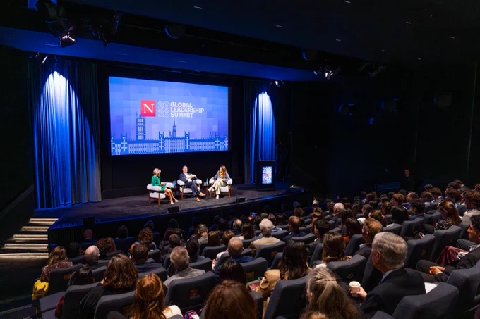 Three people on stage. Two panelists, Dame Emma Walmsley (Former CEO, GSK) and Chistopher Viehbacher (President and CEO, Biogen) sit with Joseph E. Aoun (President, Northeastern) who moderates the converstaion. They sit on stage in a dark auditorium, facing an audience. A large screen behind them displays \"2026 GLOBAL LEADERSHIP SUMMIT\" over a blue cityscape. The audience of adults fills rows of dark seats, attentively watching the discussion.