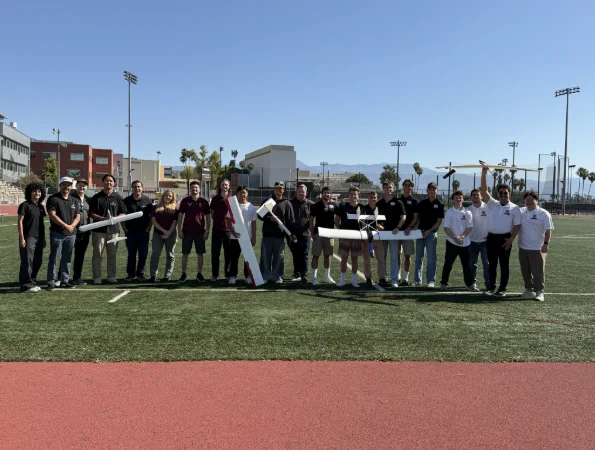 A group of people standing on a sports field under a clear blue sky. They are holding two large model planes. The surroundings include buildings, palm trees, and mountains in the distance.