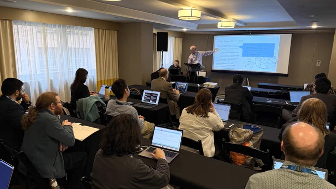 A photograph of the tutorial session, from the back of the room. Two people are at the front of the room, facing the audience and giving the tutorial presentation. About a dozen audience attendees are seated at tables, many with laptops open.