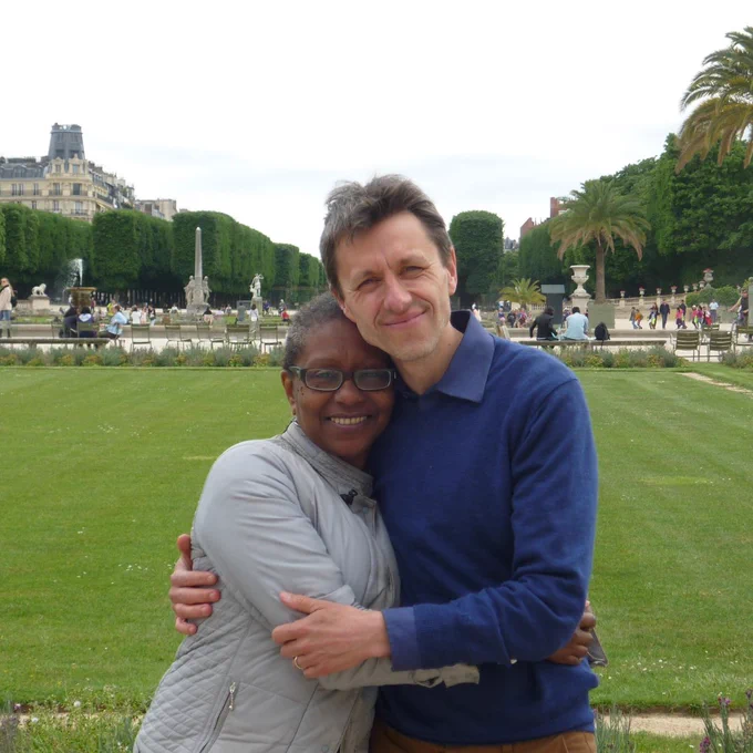 James and his wife Maria embrace. They are standing outside in a park, with foliage and trees behind them.