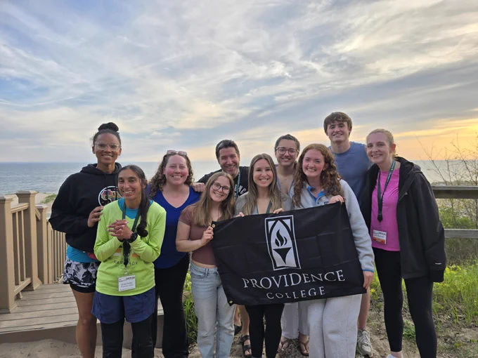 A group of individuals is smiling and standing outdoors near a wooden railing with the ocean in the background. They are holding a black banner with the "Providence College" logo and text. The sky is partially cloudy with a hint of sunset.