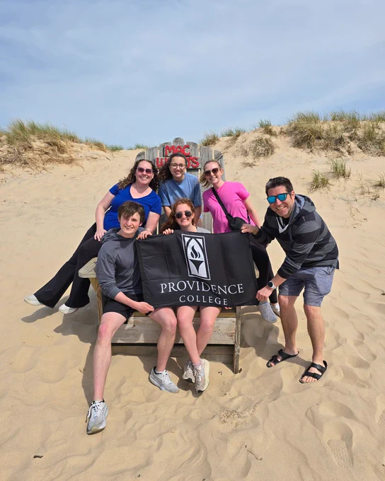 A group of six people are sitting together on a wooden structure in a sandy area with grass-covered dunes in the background. They are holding a black flag with the "Providence College" logo and text. The sky is partly cloudy.