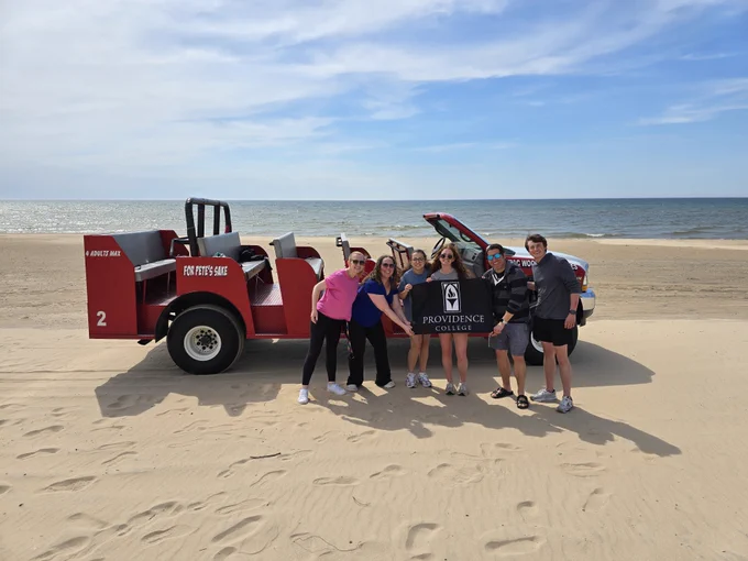 A group of people stands on a sandy beach beside a red Zamboni with "Providence College" displayed on it. The ocean and a clear sky are in the background.
