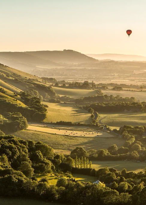 A single hot‑air balloon floats above the gently rolling green hills of the South Downs under a clear, open sky.