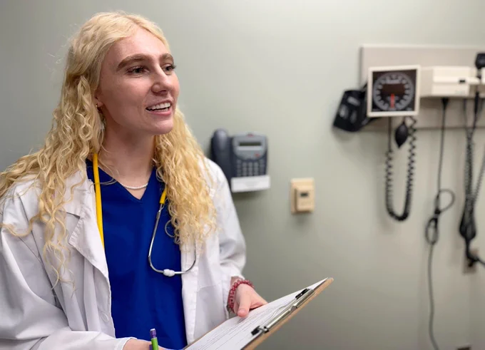 A smiling medical professional with curly blonde hair holds a clipboard in a clinic room. She wears a stethoscope, conveying a welcoming tone.