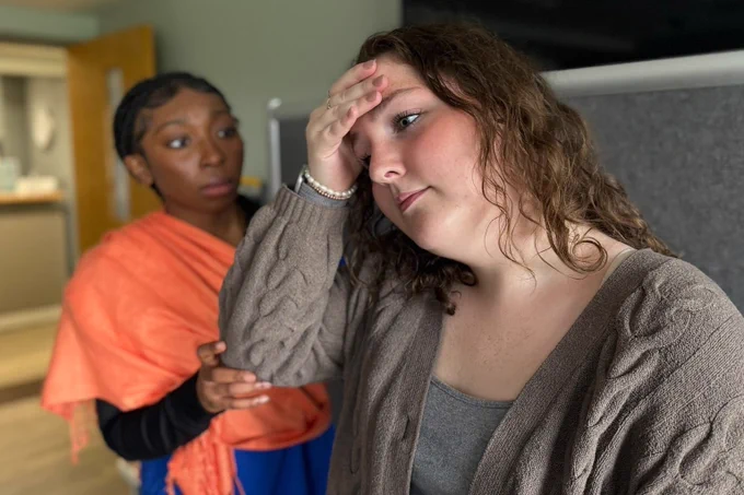 Two women are standing in an indoor setting. One looks concerned with her hand on her forehead, while the other gently touches her shoulder, offering comfort.