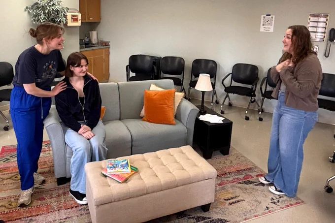 Three actors perform a dramatic scene in a waiting room setting with a grey couch, tufted ottoman, and black office chairs. One woman stands on the right looking emotionally touched, while two women on the left interact on and around a sofa.