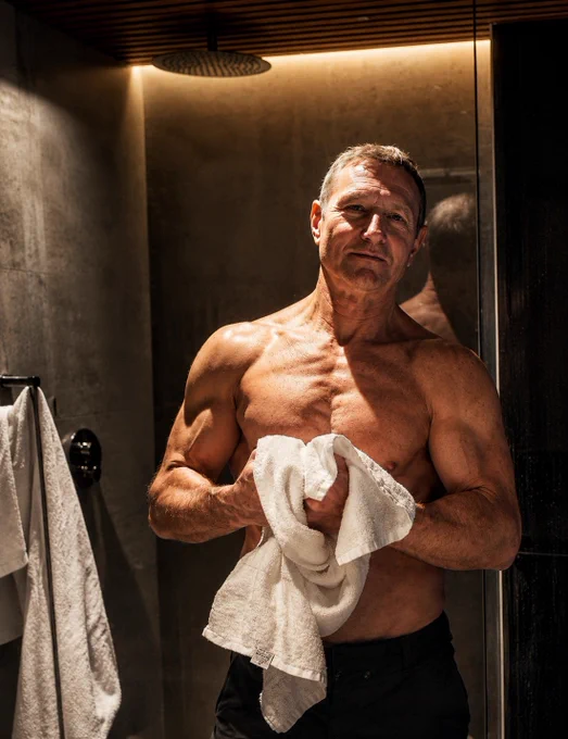 Man after workout standing in a bathroom holding a towel, showing a fit physique at 63. A quiet post-training moment focused on healthy aging, longevity, fitness after 60, and staying active.


