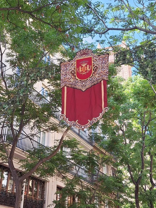 Estandarte rojo con escudo ornamentado colgado entre árboles frente a un edificio con balcones.