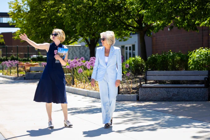 On a sunny campus walk, a blonde woman in a navy dress points, holding a \"CAMPUS TOUR\" brochure. Another woman in a light blue pantsuit walks next to her, smiling. Green trees, purple flowers, and buildings frame the paved pathway.