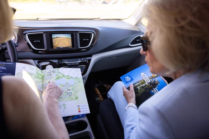 Two people in a car. The person on the left points at a map with a legend: Existing, New, Proposed building projects. The person on the right holds a brochure displaying the \"University at Buffalo\" logo and buildings. The car's dashboard screen shows a rearview camera view of a parking lot.