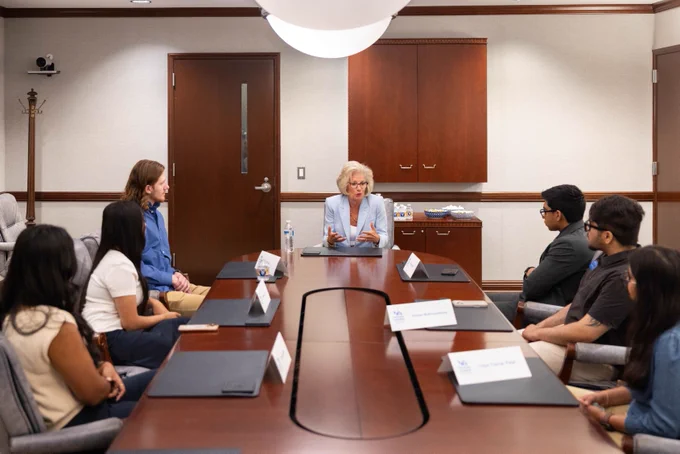 A group of five young people and one woman in a light blue blazer are seated around a U-shaped conference table. The woman is speaking and gesturing, while the others listen attentively.