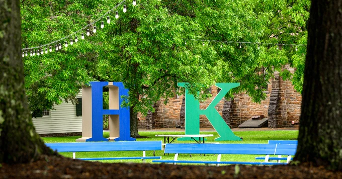A vibrant outdoor scene featuring large, colorful letters "H" and "K" on a grassy area. String lights hang above, with trees providing a lush green backdrop and benches scattered around.