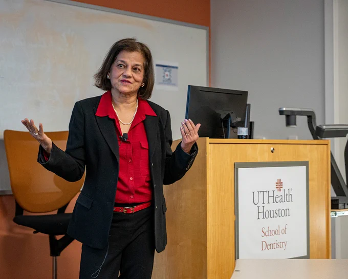 A person stands beside a podium featuring the UTHealth Houston School of Dentistry logo, wearing a black suit with a red shirt. They are speaking, with hands raised in a gesture. A computer monitor sits on the podium. Behind them is a whiteboard and an orange chair in a classroom setting.
